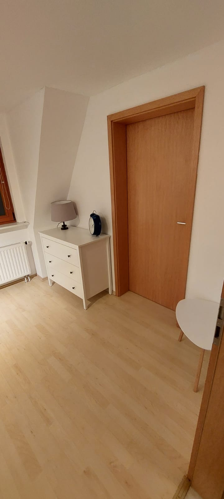 A bright room features light wooden flooring and a warm wooden door. A small white chest of drawers is positioned next to a bedside lamp. A white, round side table is placed in the corner, while a radiator is visible beneath a window.