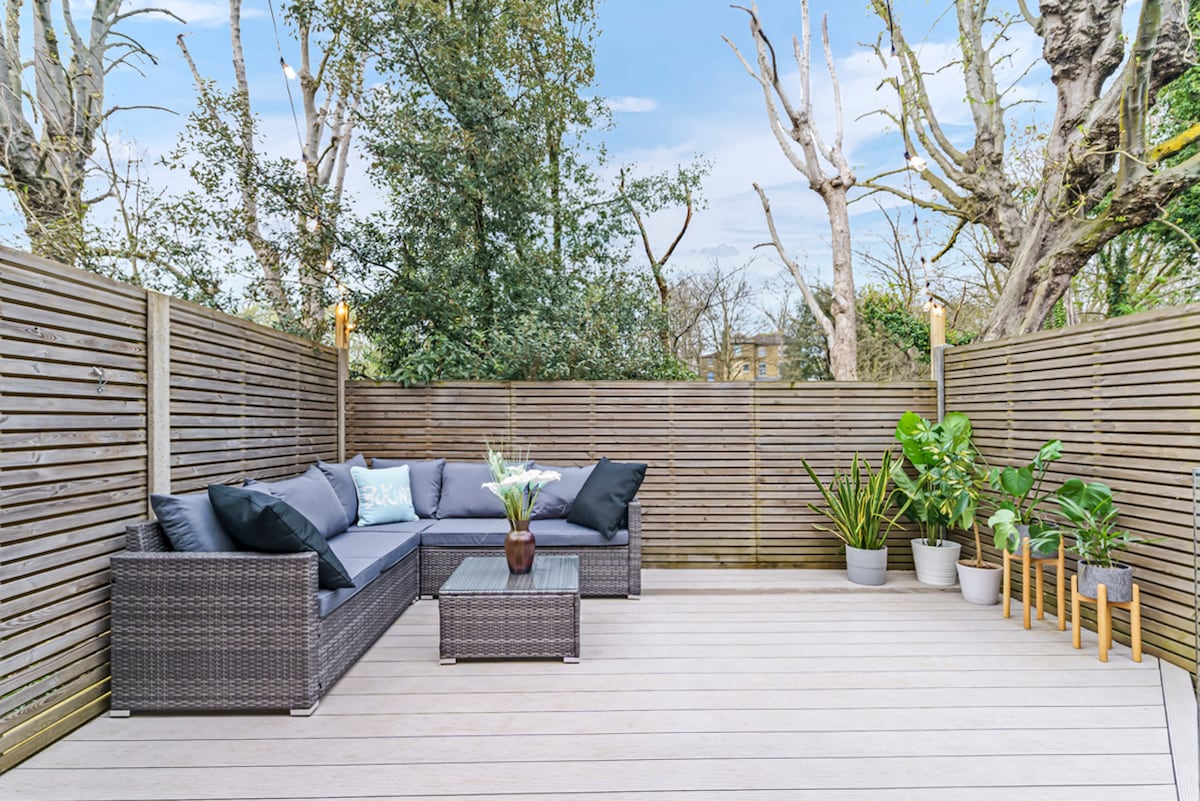 A serene outdoor decking area features comfortable seating arrangements with plush cushions. The space is surrounded by wooden fencing and lush greenery from nearby trees. Potted plants add a touch of nature, creating an inviting atmosphere for relaxation.