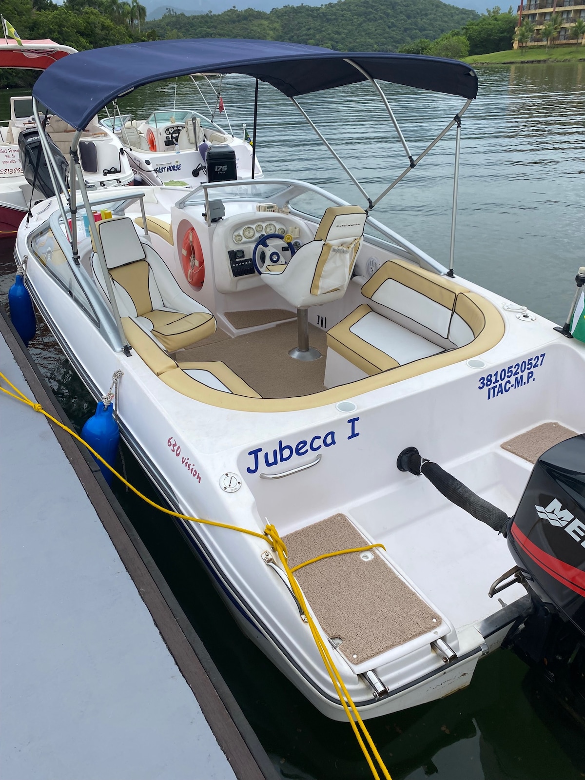 A comfortable boat named Jubeca I is docked alongside a calm body of water. The interior features padded seating in a light tan and cream color scheme. A steering console, equipped with various controls, is visible at the helm, offering a spacious area for relaxation.