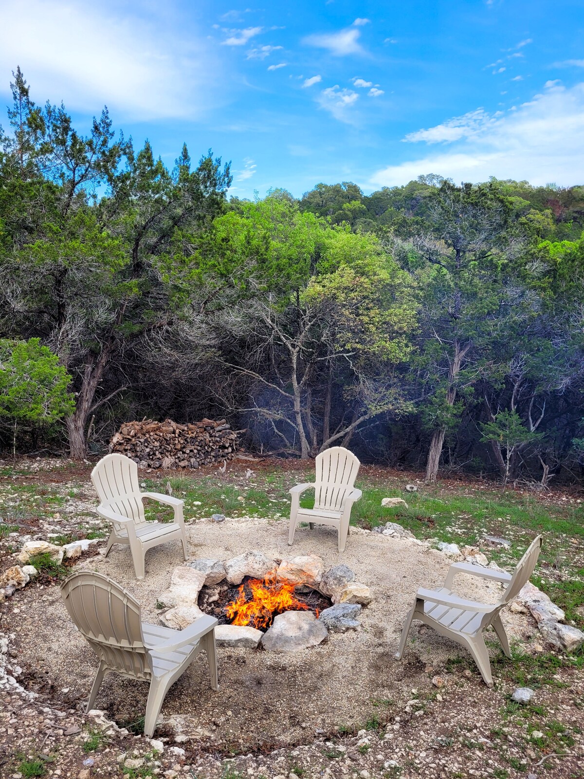 A fire pit area is surrounded by four light-colored chairs, set among a natural landscape of trees. Rocks are arranged to form the pit, with a small fire burning in the center. A stack of firewood is visible in the background.