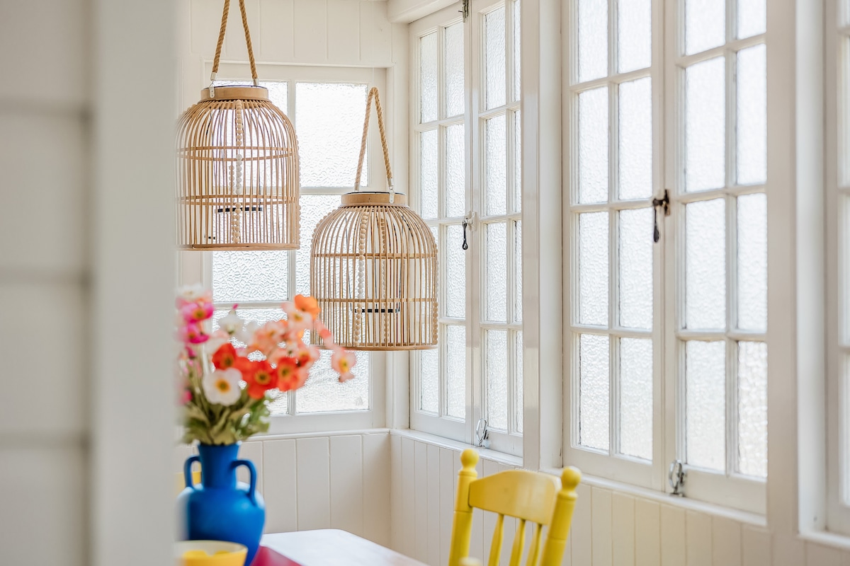 A dining area is filled with natural light from large windows, showcasing two woven pendant lights hanging from the ceiling. A vibrant bouquet of flowers sits in a blue vase on the table, complemented by a bright yellow chair arranged nearby.