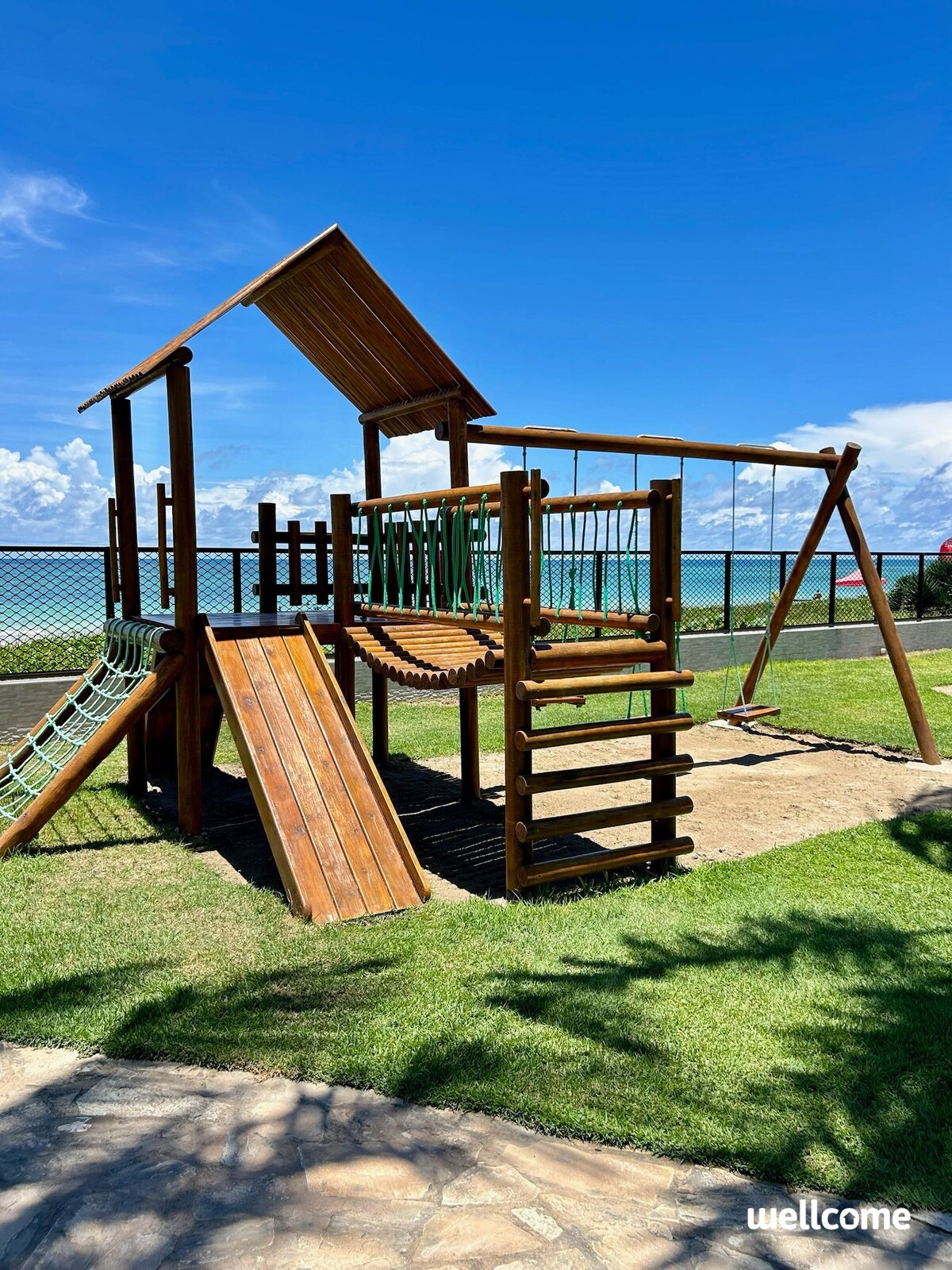 A wooden playground structure is situated on a grassy area, featuring a slide, climbing elements, and swings. The background offers a view of the ocean under a clear blue sky, enhancing the outdoor play environment.