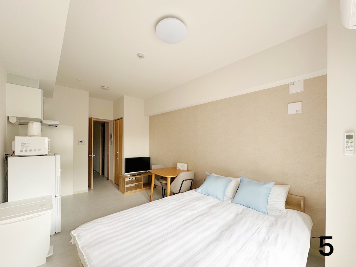 A well-lit room features a spacious bed with crisp white bedding and two blue pillows. An adjacent kitchen area includes a refrigerator and microwave, while a dining table with a chair is positioned nearby. A television is also present, enhancing the room's functionality.