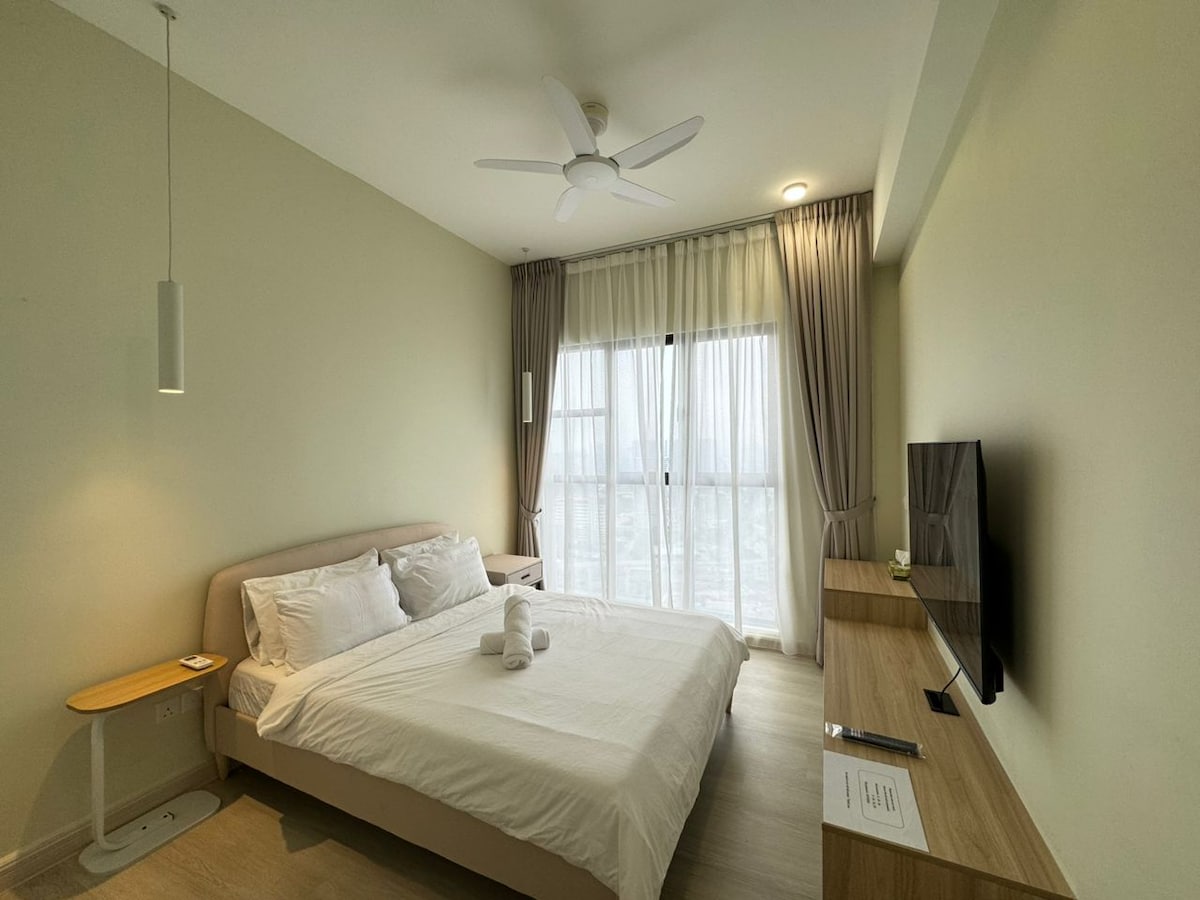 A spacious sleeping area contains a cozy bed with sanitized linens and pillows, accentuated by soft natural light filtering through large windows. A minimalist wooden desk and TV add functionality, while a ceiling fan provides ventilation, contributing to a comfortable ambiance.