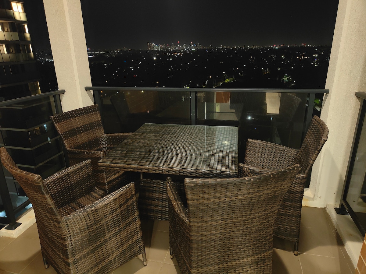 An outdoor dining space is presented with a glass-top table surrounded by six wicker chairs. The balcony offers expansive views of the city skyline against a night sky, with twinkling lights illuminating the distant horizon.