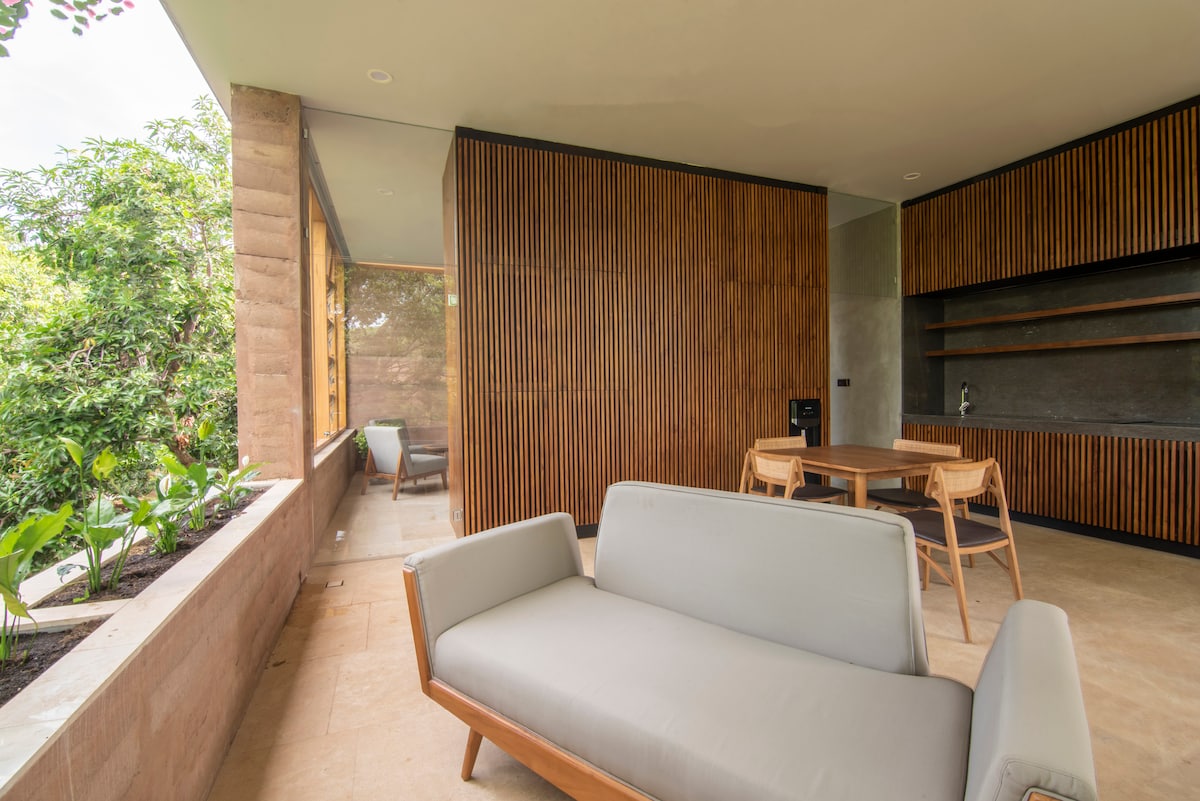 A modern outdoor living space features a light grey sofa positioned near large windows, allowing views of the surrounding garden. A wooden dining table with light-colored chairs occupies the area, while natural elements like plants and rammed earth walls create a seamless connection to nature.