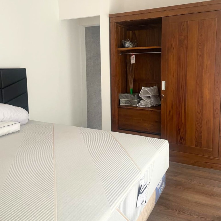A spacious bedroom is presented with a neatly made bed featuring a light-colored fitted sheet. A wooden wardrobe stands closed beside the bed, providing storage space. The minimalistic design allows for easy movement and comfort.