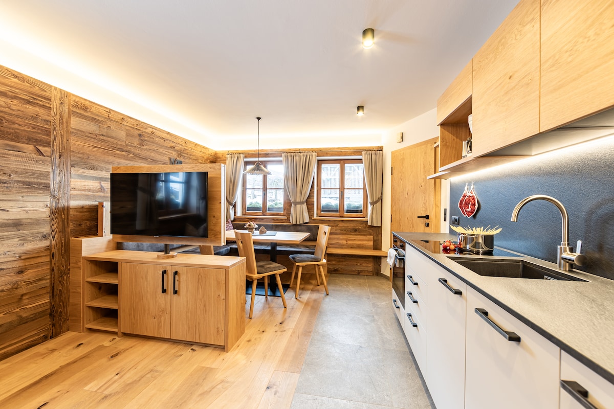 The modern kitchen features a spacious countertop and sleek cabinetry. A dining area with wooden chairs is positioned next to a large window, allowing natural light to fill the space. A flat-screen TV is mounted on the wooden entertainment unit, blending with the warm wood accents.