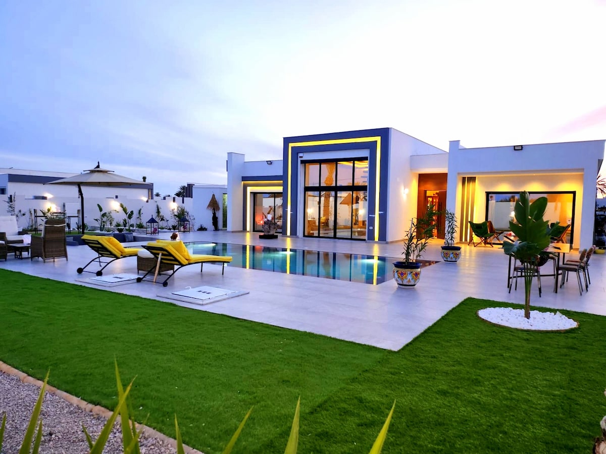 The villa features a modern architectural design with expansive glass doors and a swimming pool surrounded by a well-kept lawn. Comfortable lounge chairs are positioned poolside, while decorative pots and outdoor furniture create a welcoming atmosphere for relaxation.