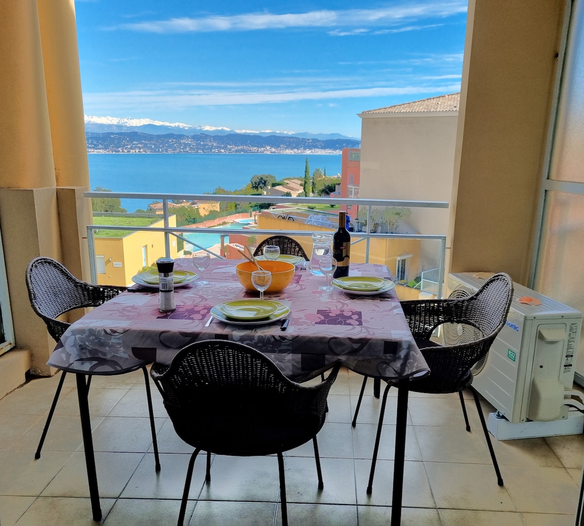 A dining table set for four is positioned on a terrace overlooking panoramic sea views and snow-capped mountains. Four black wicker chairs surround the table, which features a decorative tablecloth and an array of tableware, complemented by a bottle of wine and a bowl.