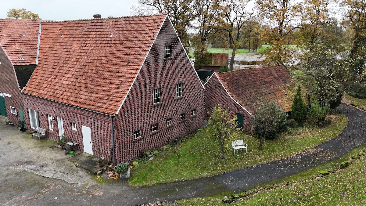 An expansive view of the historic farmhouse showcases its red brick exterior and traditional triangular roof. Surrounding greenery includes a gravel path leading to a small garden area, enhanced by seasonal foliage. The quiet countryside setting reflects a serene ambiance.