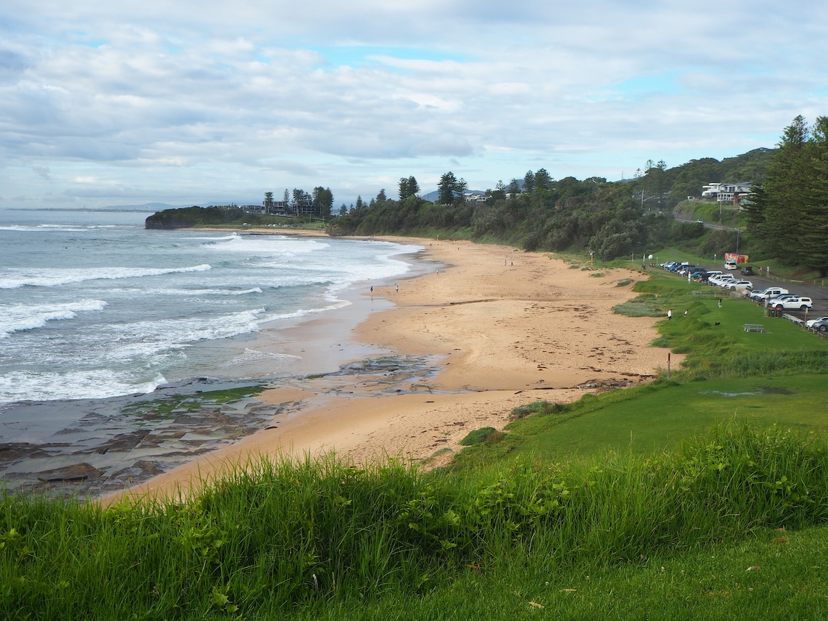 A sandy beach stretches along the coastline, bordered by gentle waves and rocky patches. A grassy area separates the beach from the view, while palm trees and distant buildings frame the shoreline. A few people can be seen enjoying the seaside in the background.