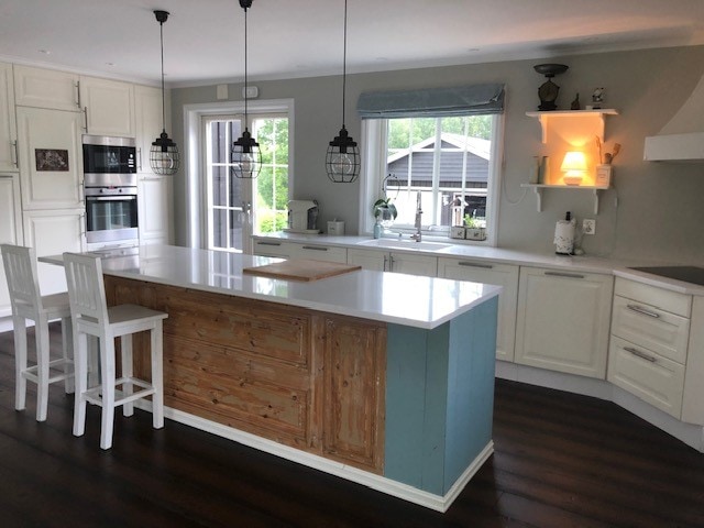 A modern kitchen features a large central island with a light wood finish, complemented by white cabinetry. Pendant lights hang above, illuminating the space. An abundance of natural light enters through sliding glass doors, with a view of the serene outdoor area providing a backdrop.