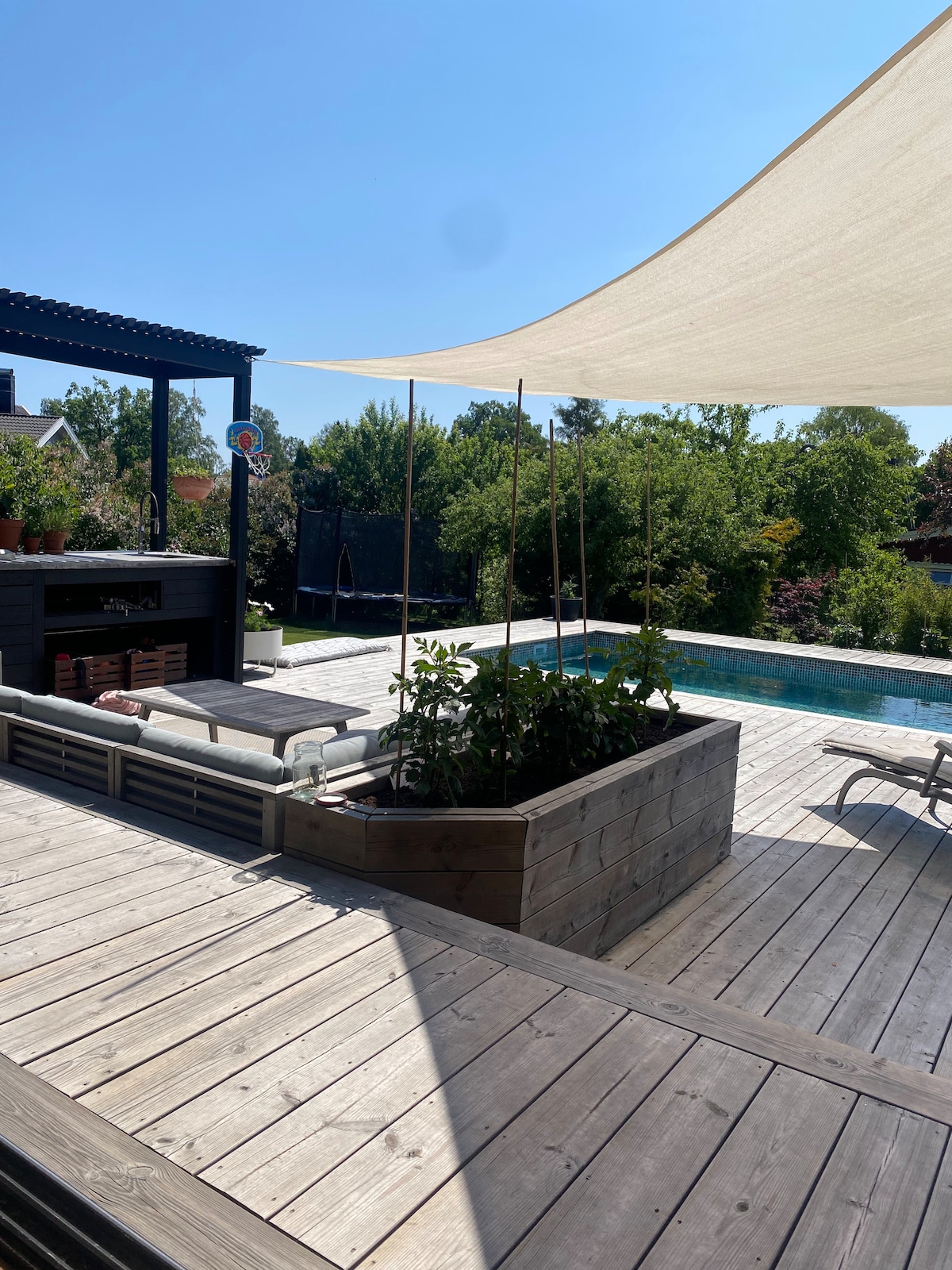 An outdoor patio is featured with wooden decking, a shaded area provided by a canopy, and a built-in outdoor kitchen. A modern swimming pool is visible in the background, surrounded by lush greenery, adding to the serene setting.