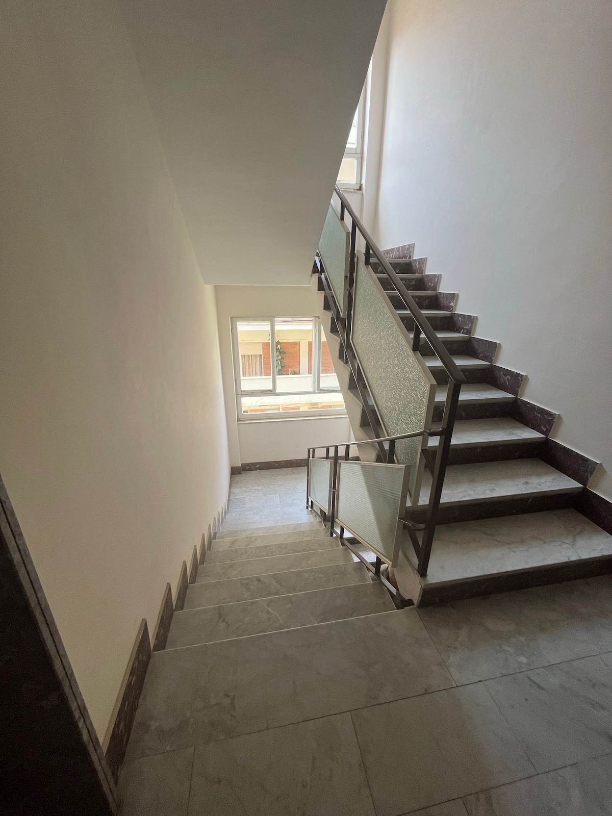 A staircase leads upward, featuring smooth marble steps. Natural light enters from a nearby window, illuminating the neutral-colored walls. A glass railing runs alongside, providing a modern touch to the space.