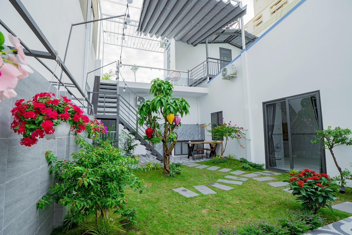 A serene courtyard features a lush green lawn surrounded by vibrant flowers and a small tree. Paths of stepping stones lead through the garden. The area is enclosed by two-story buildings with stairs visible in the background, creating an inviting space for relaxation.