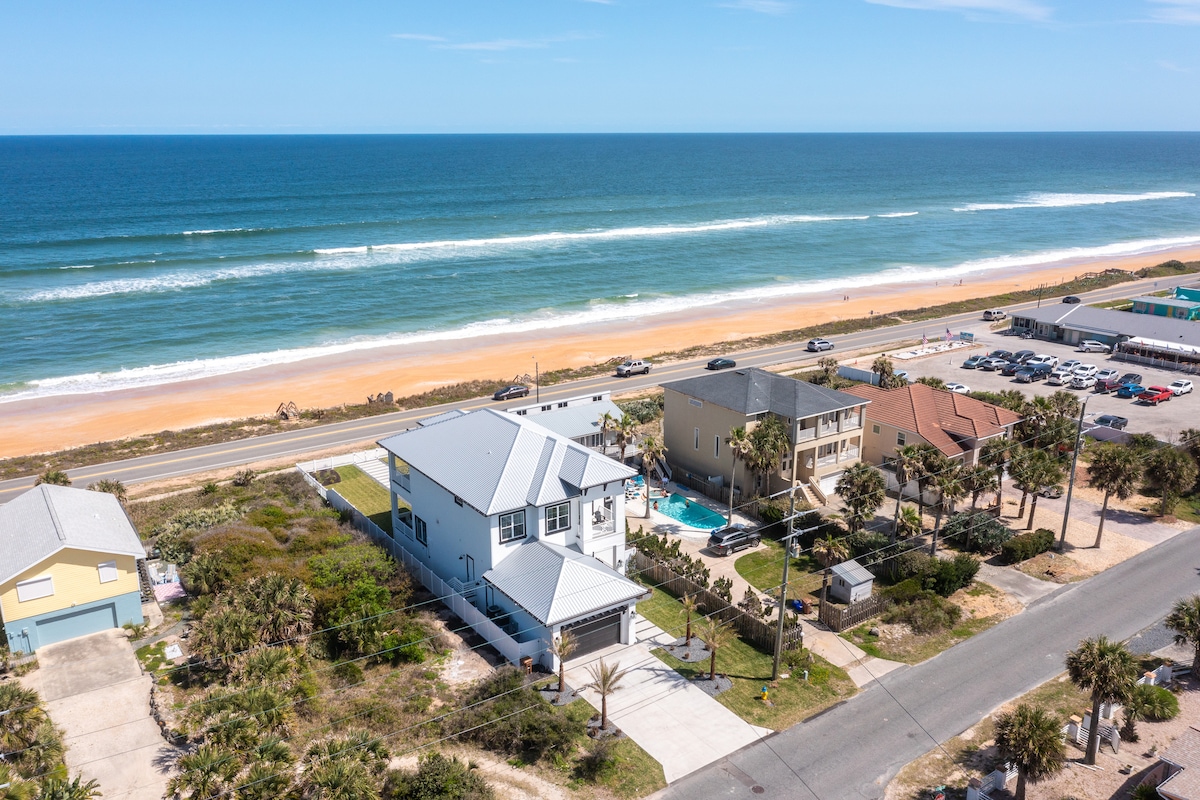 Aerial view captures a spacious beachfront home adjacent to sandy shores. Bright ocean waves gently lap the beach. The home's modern architecture stands out against the backdrop of the coastline, while palm trees and surrounding residences provide a coastal community feel.