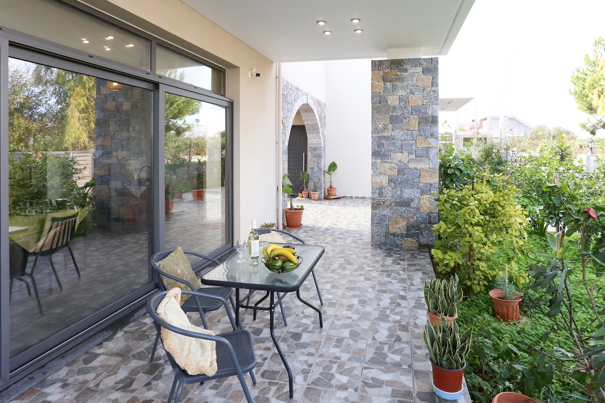 A covered terrace is visible with modern outdoor furniture, including a glass table and chairs. The space is bordered by a well-maintained garden featuring potted plants and greenery, creating an inviting area for relaxation.