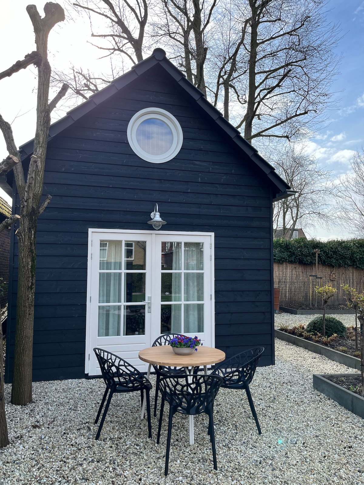 An exterior view of a charming black wooden casita is displayed, featuring large glass doors that open to the outside. A round table with four black chairs is positioned in front, surrounded by a gravel yard and greenery, enhancing the serene environment.