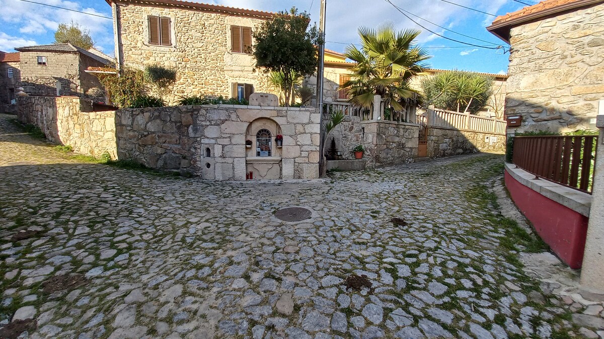 A cobblestone path leads to a small, stone fountain surrounded by greenery and traditional buildings with stone facades. Visible are two windows on the nearby structure, framed by a charming garden area with a palm tree, contributing to the rustic village ambiance.