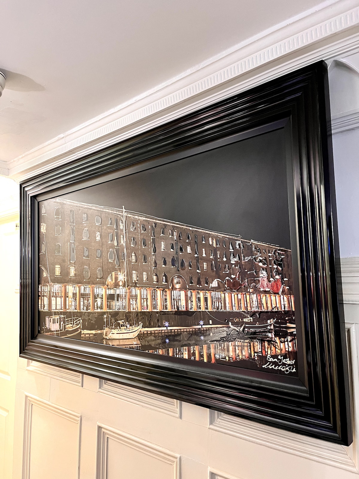 A large framed painting hangs on the wall, depicting a nighttime scene of a waterfront with historic buildings, boats, and reflections in the water. The black frame complements the artwork, enhancing its visual impact.