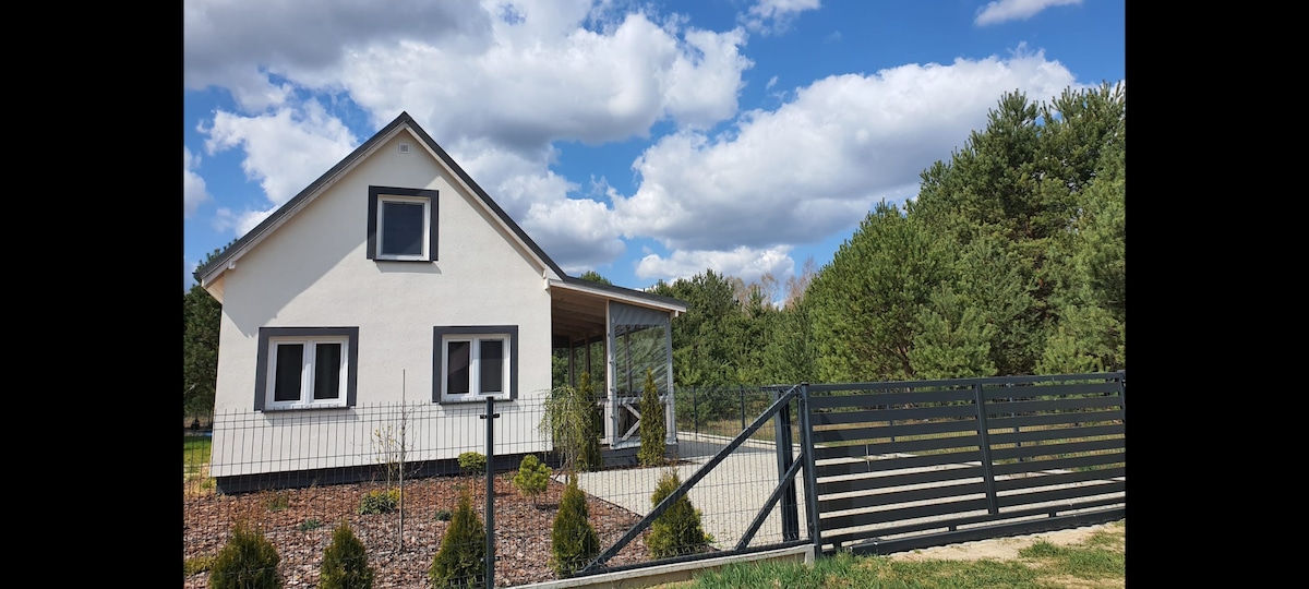 A charming house is presented with a light exterior, featuring a covered porch and large windows. The property is fenced, with neatly arranged shrubs and greenery visible in the surrounding landscape. A clear sky with clouds adds to the serene outdoor ambiance.