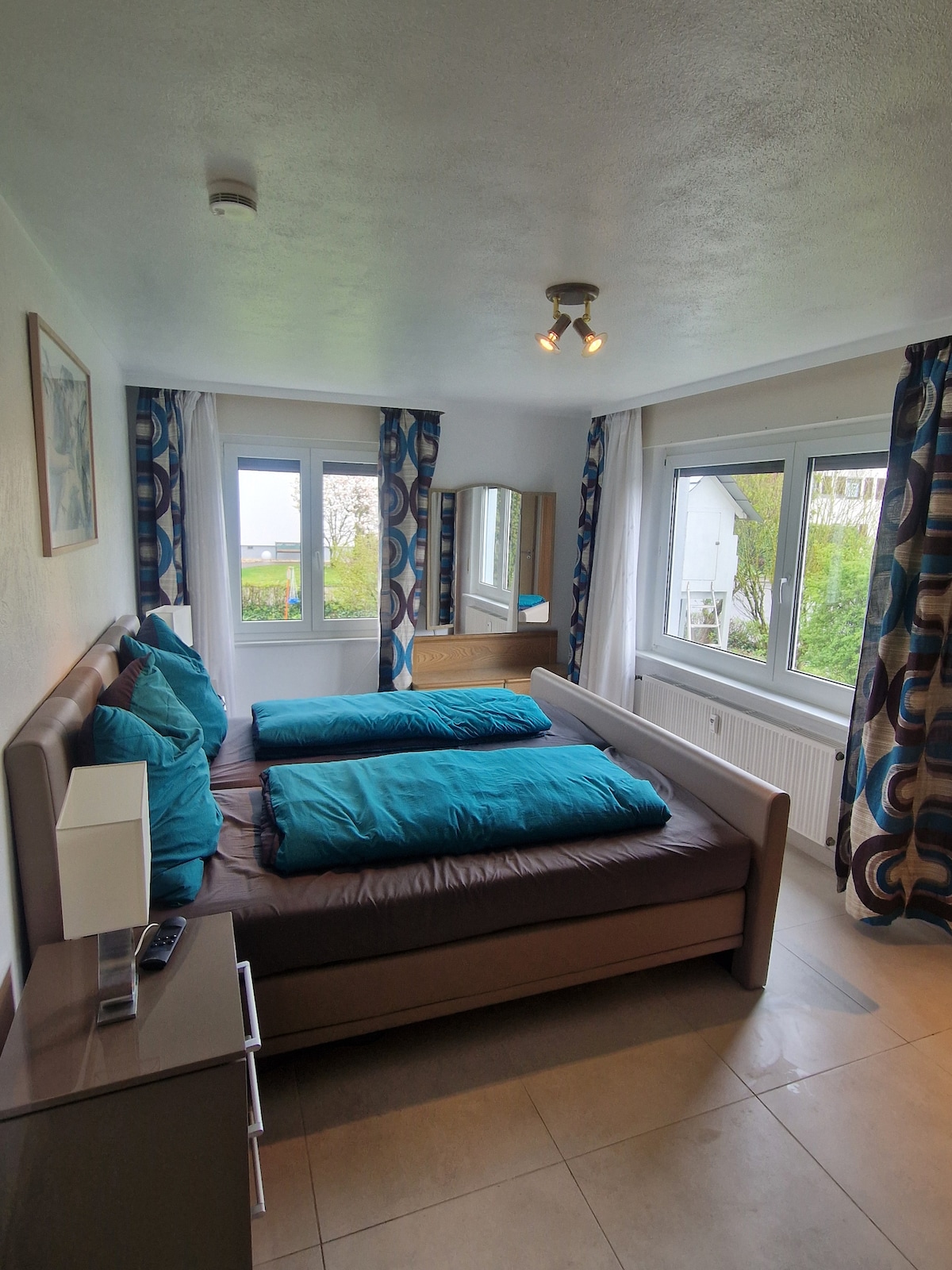 A well-lit bedroom features a spacious bed with two aqua-colored duvets and pillows. Large windows allow natural light to fill the room, while patterned curtains frame the view. A bedside table with a lamp and a dresser provide functional storage.