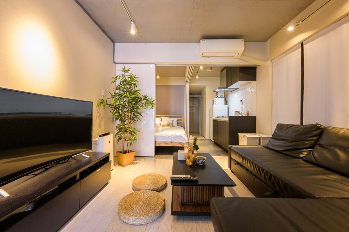 A spacious living area features a sleek black sofa and a low wooden coffee table adorned with decorative items. A large television is positioned against a wall, while a lush plant adds a touch of greenery. A bed can be seen through a partition, creating a cozy atmosphere.