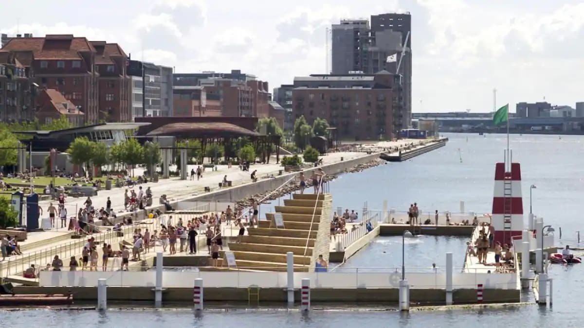 The image captures a vibrant waterfront scene at Islands Brygge, showcasing the popular harbour bath with visitors enjoying the water. Lush greenery lines the promenade, complemented by modern structures in the background. Bright sunny skies and reflections on the water enhance the lively atmosphere.