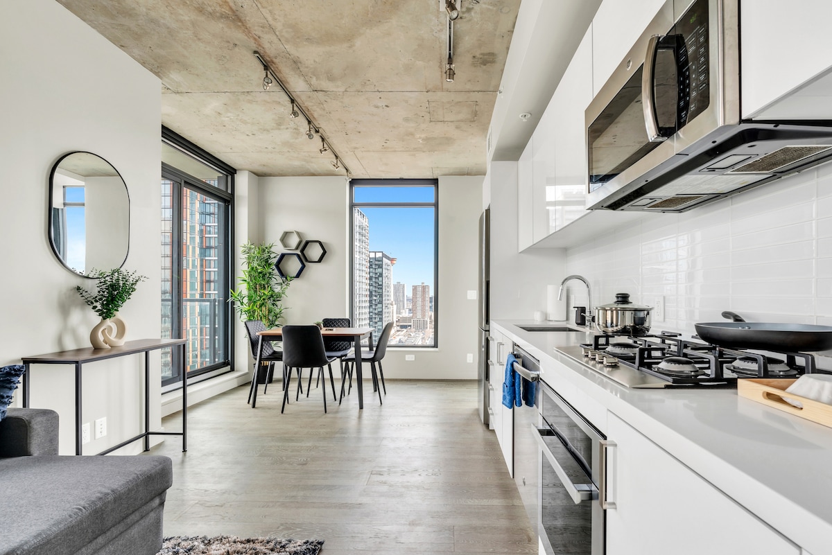 The open-concept living area includes a well-equipped kitchen featuring modern appliances and sleek cabinetry. A dining table for four is positioned near a window, showcasing city views. Natural light floods the space, highlighting the contemporary decor and inviting indoor plants.