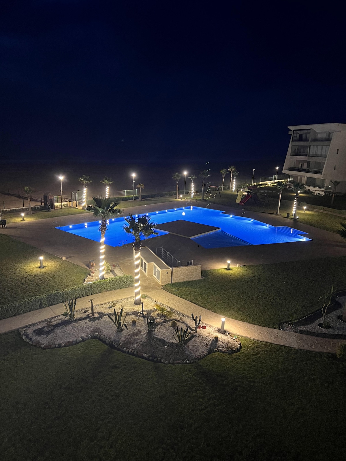 At night, the illuminated pool area presents a vibrant blue color, surrounded by palm trees and landscaped gardens. Soft lighting enhances the outdoor space, while lounge areas invite relaxation. The distant ocean creates a tranquil backdrop, contributing to the peaceful atmosphere of the residential complex.