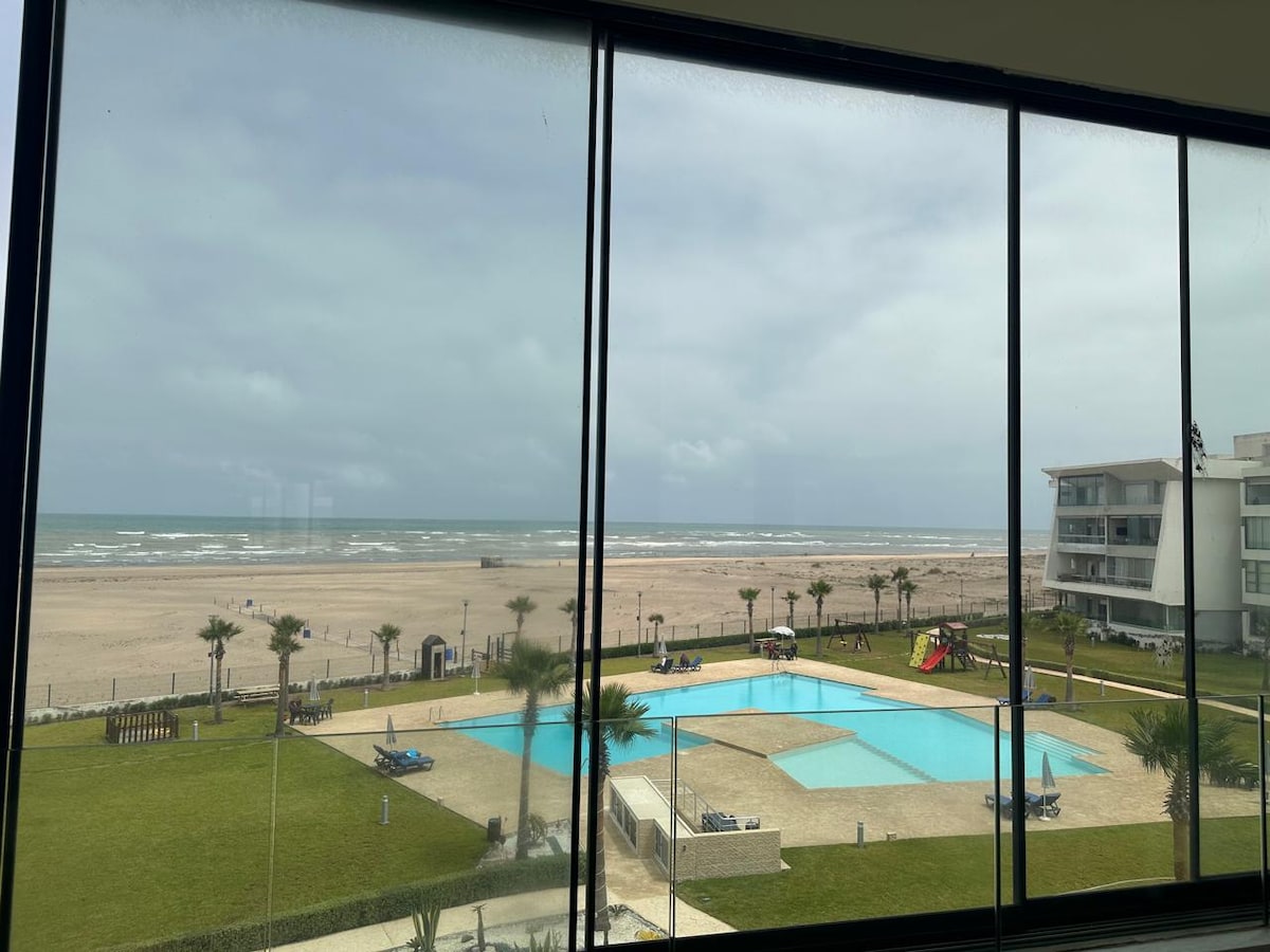 A large window offers a view of the ocean and sandy beach, framed by modern architecture. In the foreground, a swimming pool is visible, surrounded by lush grass and palm trees. A playground and pathways are also seen, creating a serene coastal environment.