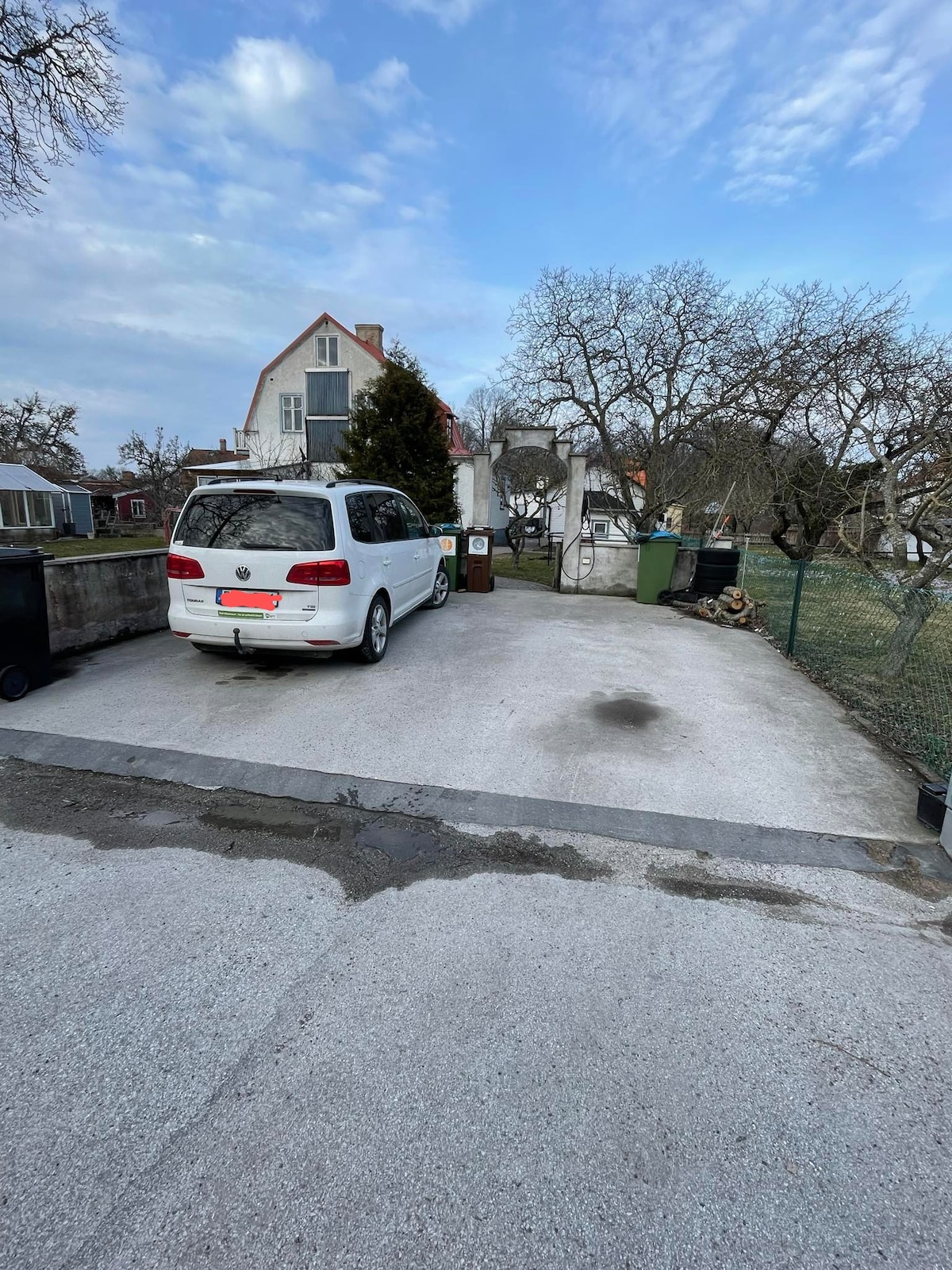 A driveway is visible leading to a white vehicle parked next to a grey concrete surface. Trash bins are placed along the edges, and a fence surrounds the area. A grassy yard and trees can be seen in the background, contributing to the outdoor scenery.