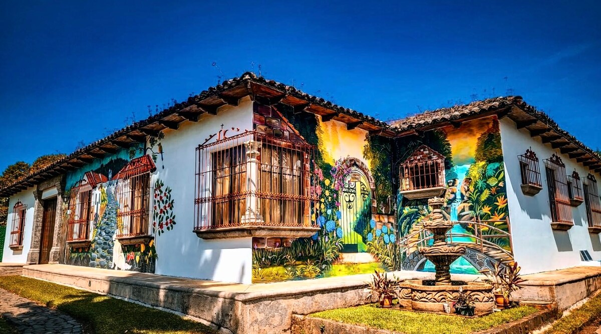 The exterior of a colorful building is adorned with intricate murals depicting nature and traditional architecture. Decorative wrought-iron windows and a central fountain enhance the inviting ambiance. Lush greenery surrounds the property, set against a clear blue sky.