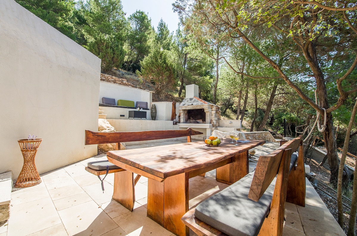 An outdoor dining area features a large wooden table and benches, surrounded by trees. A barbecue spot is visible in the background, alongside comfortable seating. Sunlight filters through the leaves, creating a serene outdoor space perfect for meals and gatherings.