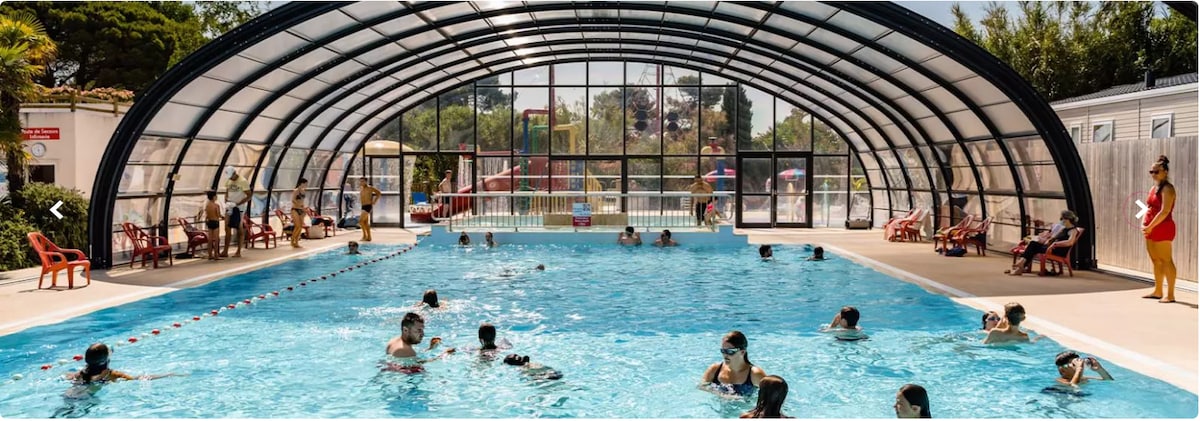 An indoor swimming pool is enclosed by a transparent arch structure, allowing natural light to fill the space. Several people are seen enjoying the water, while spacious areas for lounging with red chairs are positioned around the pool. Large glass panels provide views of an outdoor recreation area.
