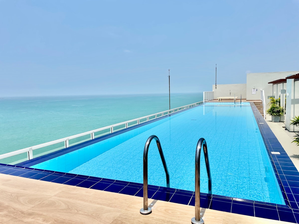 An outdoor infinity pool features clear blue water, extending towards the horizon where the ocean meets the sky. The deck is surrounded by minimalistic elements and offers unobstructed views of the serene seascape.