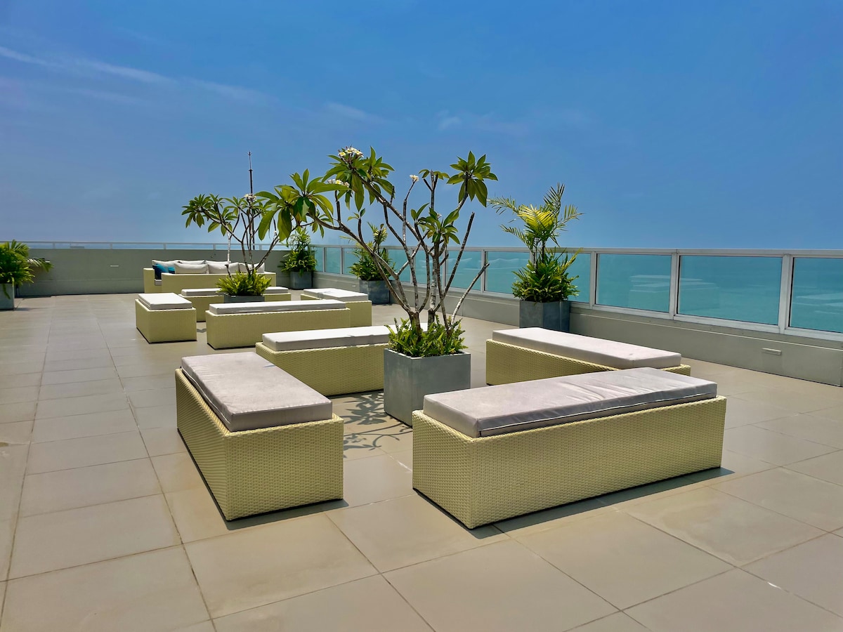 A spacious rooftop area features modern seating arrangements and planters filled with greenery. The panoramic views of the ocean are clearly visible under a bright blue sky, providing a serene environment for relaxation.