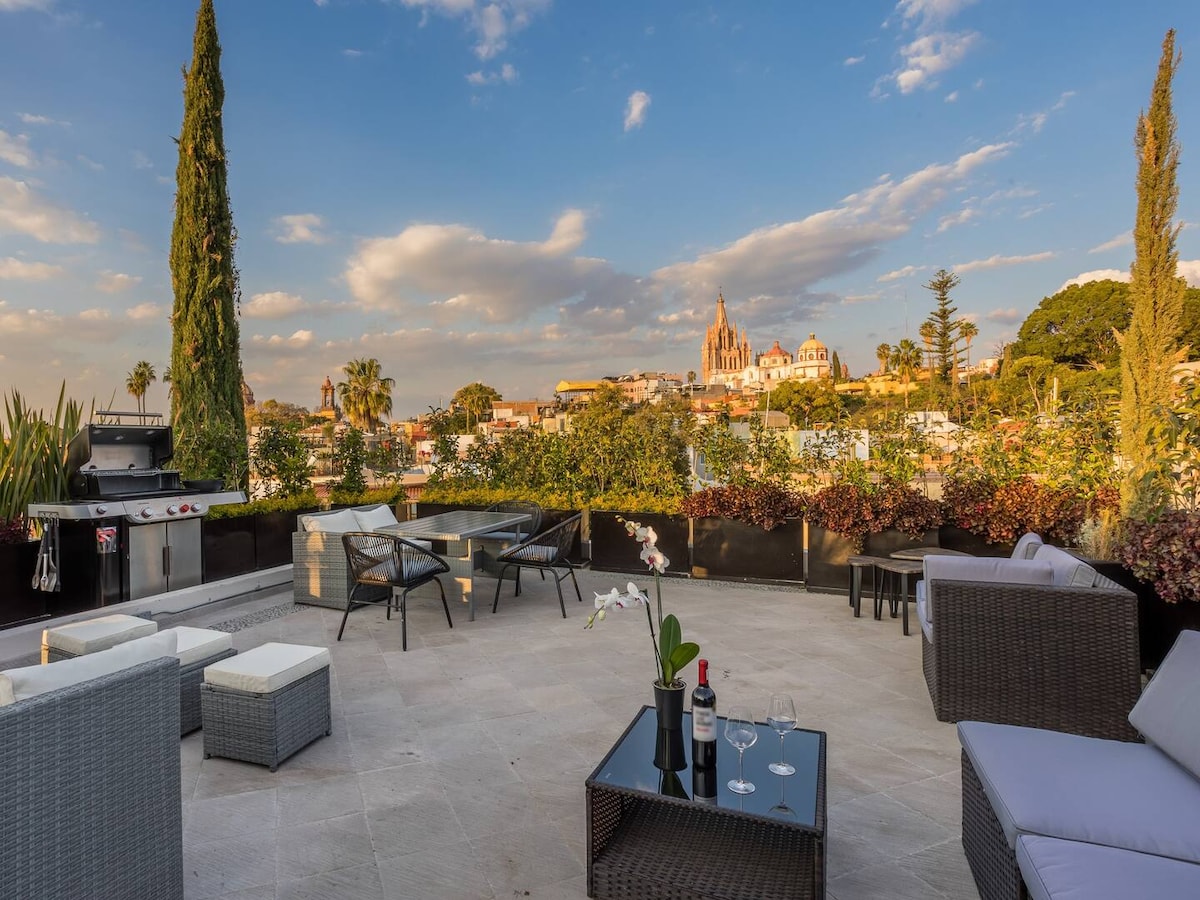 A spacious rooftop terrace is featured, furnished with comfortable lounge seating and a dining area. Lush greenery borders the space, while views of the city skyline, including notable architecture, can be seen in the distance. A barbecue grill is also present for outdoor cooking.