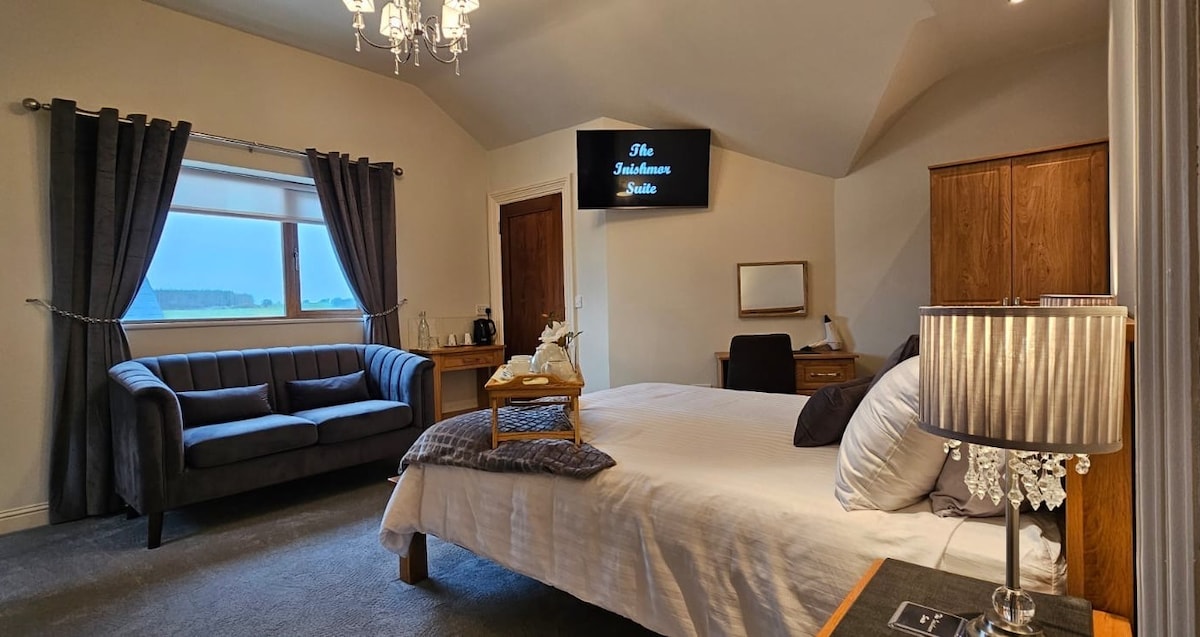 A spacious suite features a comfortable bed with crisp linens and a stylish lamp on a bedside table. A gray couch is positioned by a window, while a smart TV is mounted on the wall. Natural light fills the room, creating a cozy atmosphere.