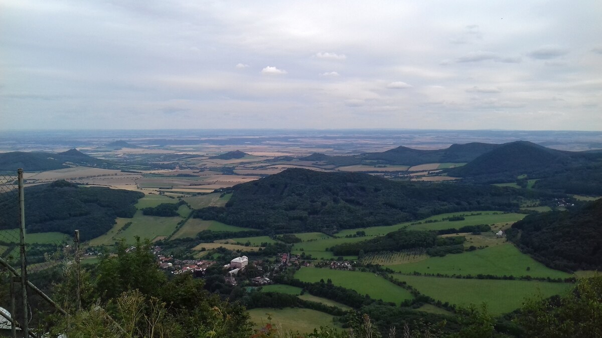 A panoramic view reveals rolling hills and expansive fields under a cloudy sky. Various shades of green characterize the landscape, with patches of forest and agricultural land visible. A small village can be seen nestled within the greenery, offering a glimpse of rural life.
