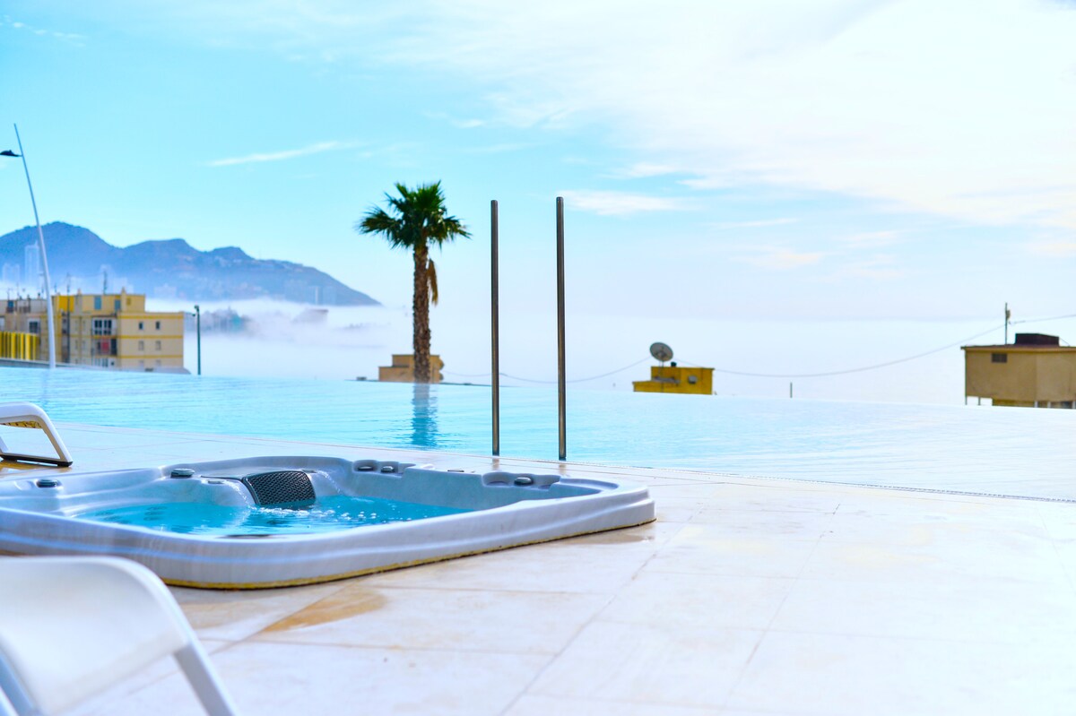 An outdoor jacuzzi is positioned near an infinity pool, overlooking a serene coastal view. A tall palm tree is visible against the backdrop of distant mountains and the expansive sky, contributing to a tranquil ambiance.