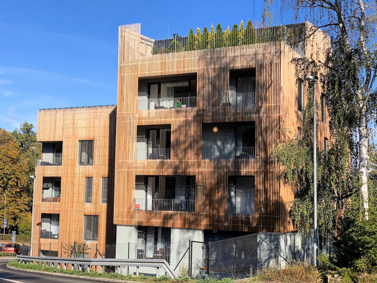 The modern building features a facade of wooden slats, blending harmoniously with the surrounding nature. Multiple balconies are visible, and the rooftop is adorned with greenery. Large windows enhance natural light, contributing to a welcoming and contemporary appearance.