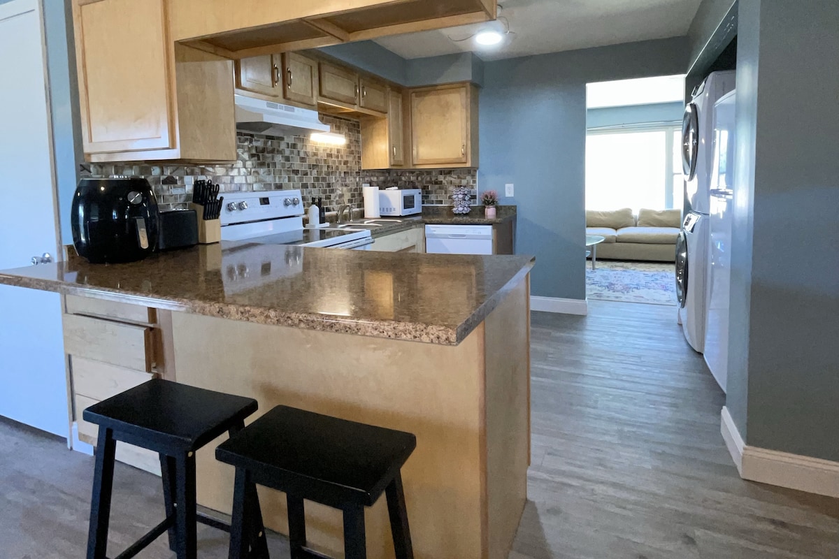 A kitchen area features honey-colored cabinetry and a granite countertop, with two black stools positioned at the island. A stove and microwave are visible, with mosaic tile backsplash adding texture. The view extends to a living area that includes a comfortable couch and natural light.