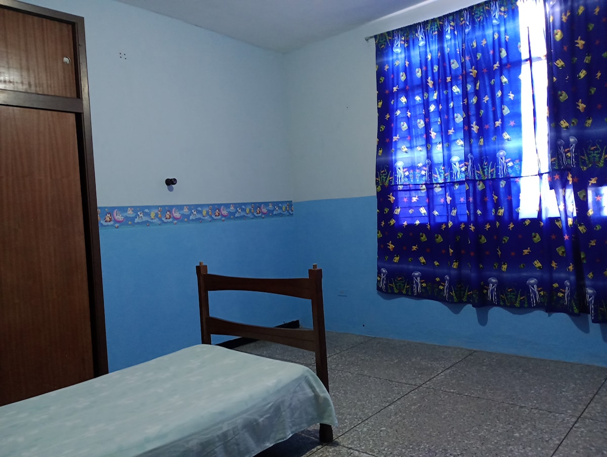 A simple bedroom features a single bed covered with a light blue sheet. A wooden headboard adds warmth to the space. Brightly patterned curtains with a marine theme frame the window, allowing natural light to illuminate the room. A closet door is visible in the corner.