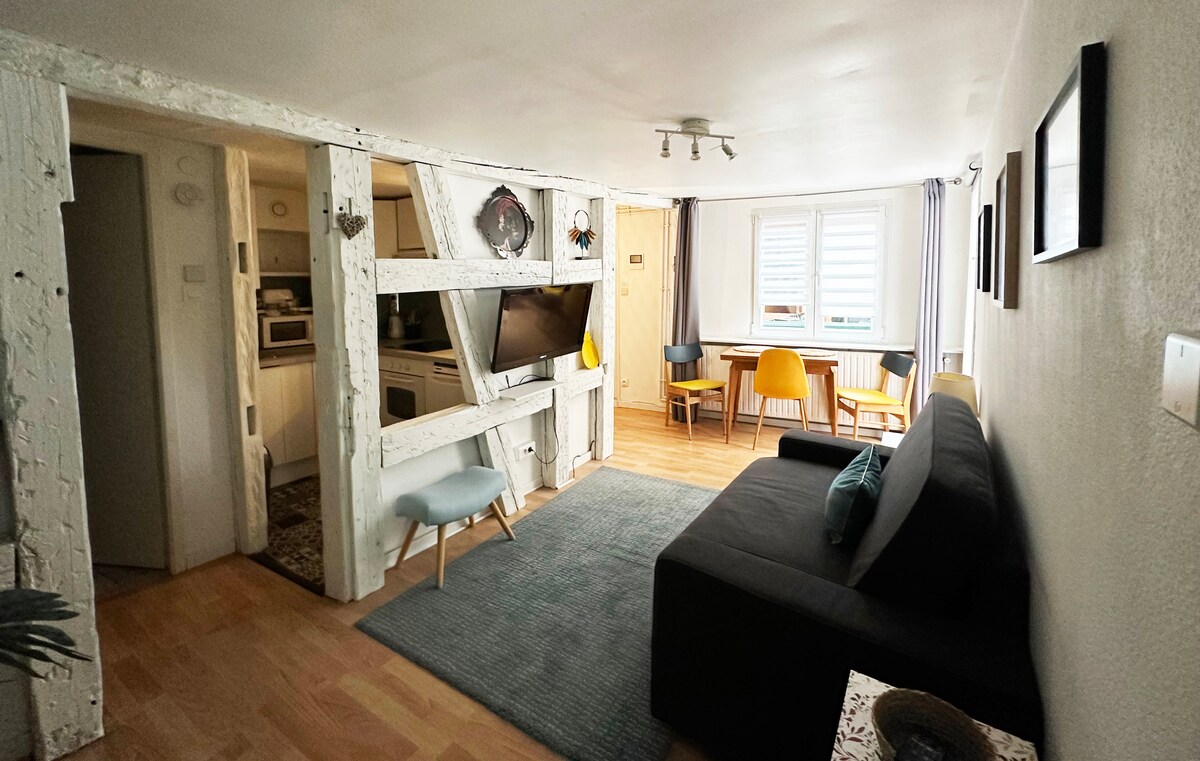 An inviting living space featuring a dark sofa and a rug on light wooden flooring. A television is mounted on a rustic white wall with wooden beams. A dining area with two chairs and a small table is visible near the window, allowing natural light to enhance the room.
