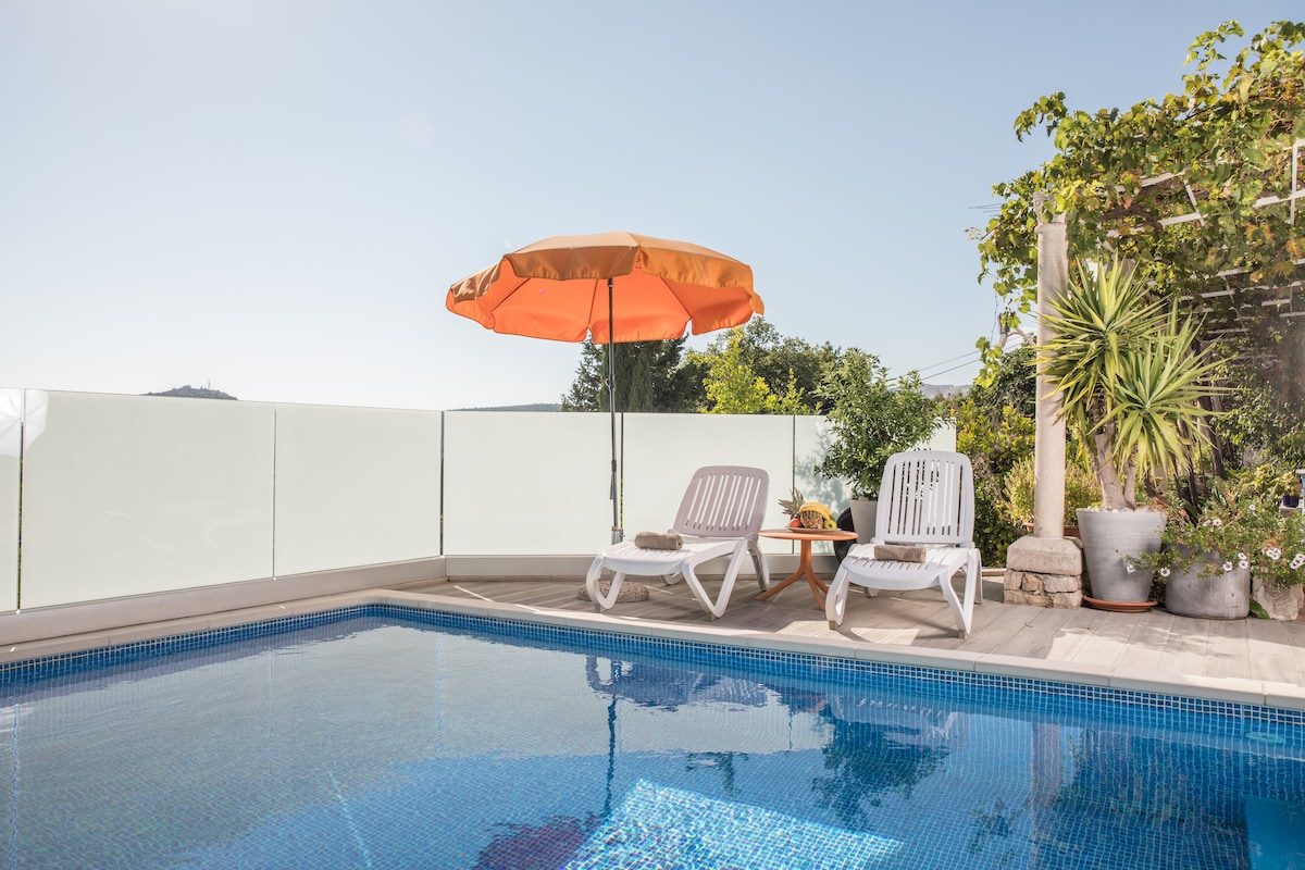 A serene swimming pool is situated in a sunlit area, bordered by clear glass walls. An orange umbrella provides shade over two white lounge chairs. Lush greenery and potted plants surround the space, contributing to the tranquil atmosphere.