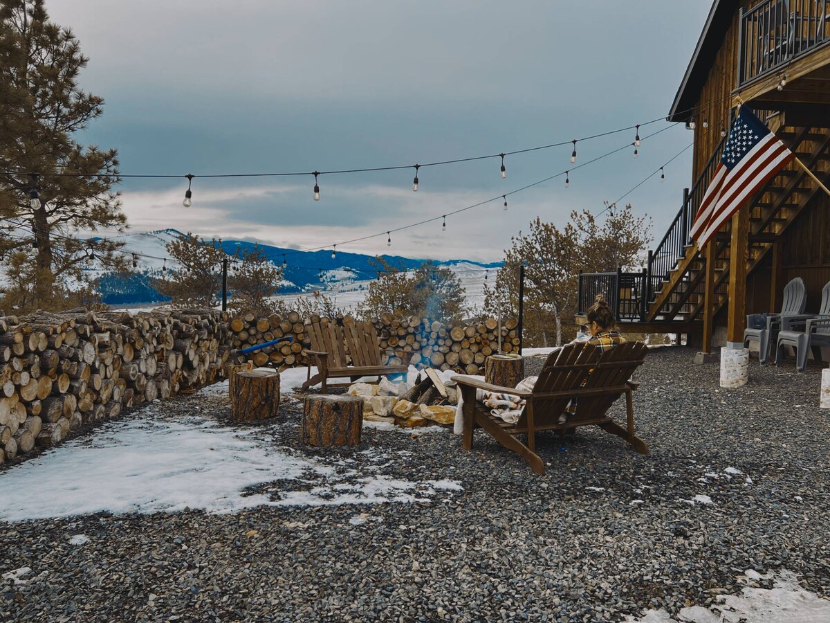 An outdoor fire pit area is surrounded by wooden seating and rustic stools. A string of lights hangs overhead, providing soft illumination against a backdrop of snow-covered ground and distant mountains. The space invites relaxation and connection under an open sky.
