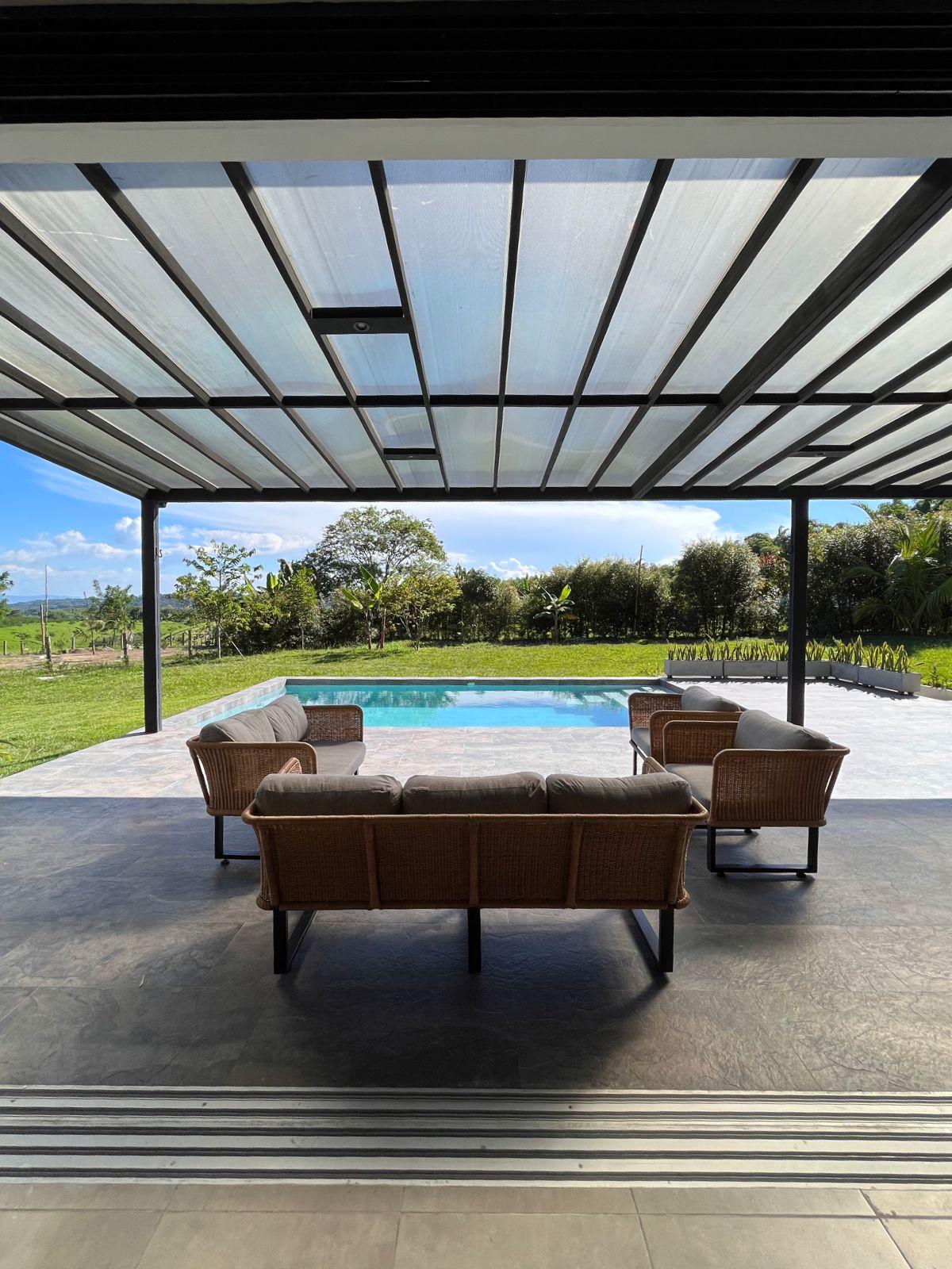 A covered terrace features a set of four rattan chairs arranged around a glass table, overlooking a private pool. Lush greenery surrounds the area, and the sky above is clear with soft clouds.
