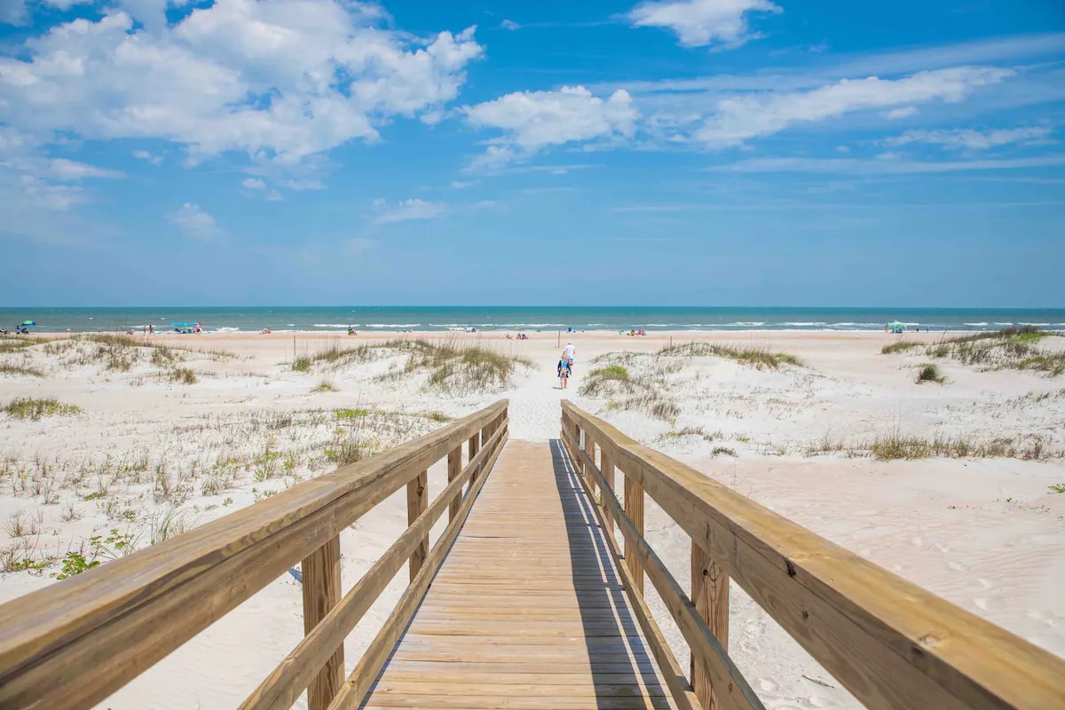 A wooden walkway leads to a serene beach, bordered by soft white sand and gentle dunes. The ocean's calm waters stretch into the distance under a clear blue sky, with a few beachgoers visible enjoying the space. Wispy clouds softly accentuate the horizon.